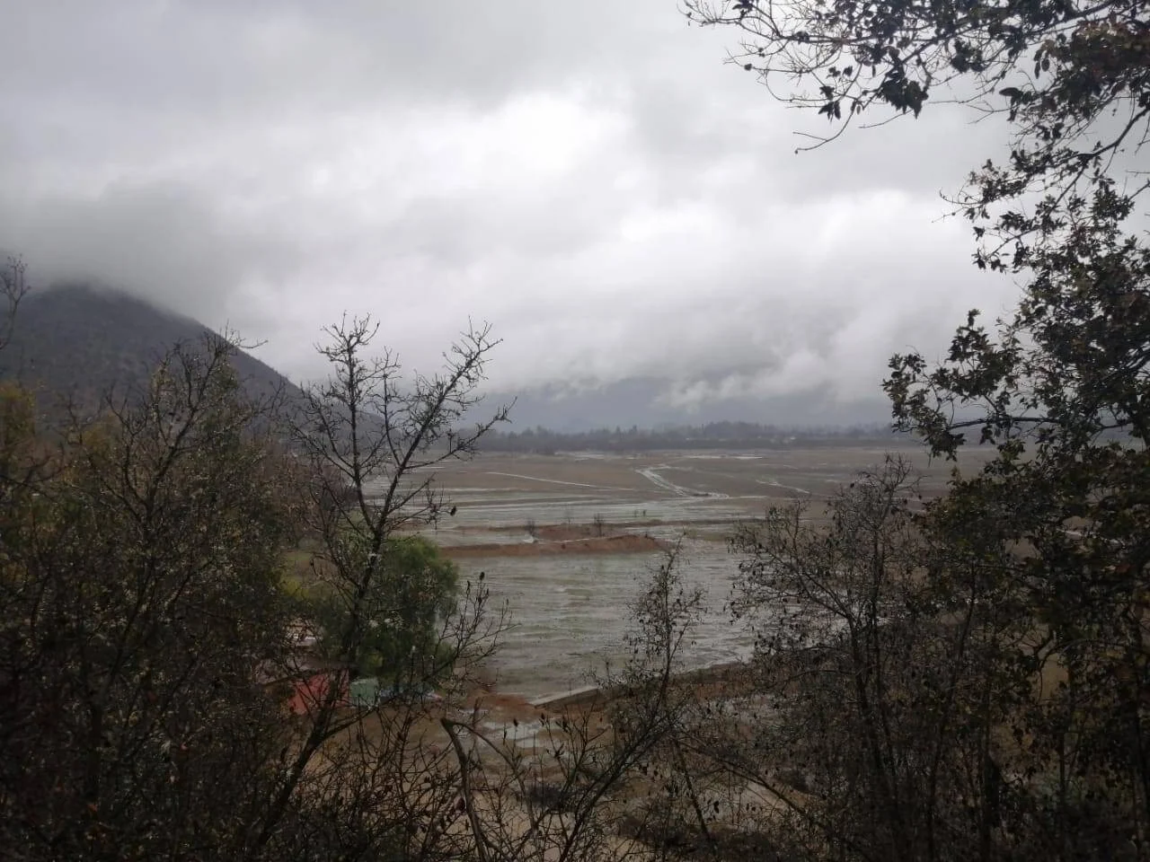 Amplio valle anegado por lluvias intensas, rodeado de cerros cubiertos de nubes bajas y vegetación sin hojas.