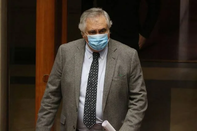Hombre mayor con traje gris y mascarilla quirúrgica azul sale por una puerta de vidrio.