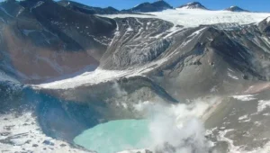 Vista aérea de un cráter volcánico con laguna turquesa, fumarolas y glaciares en las laderas de alta montaña.