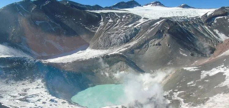 Vista aérea de un cráter volcánico con laguna turquesa, fumarolas y glaciares en las laderas de alta montaña.