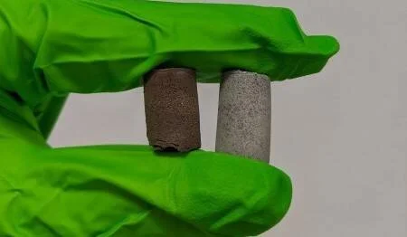 Mano con guante verde sostiene dos cilindros de material: uno marrón oscuro y otro gris claro, para comparar su composición o textura.