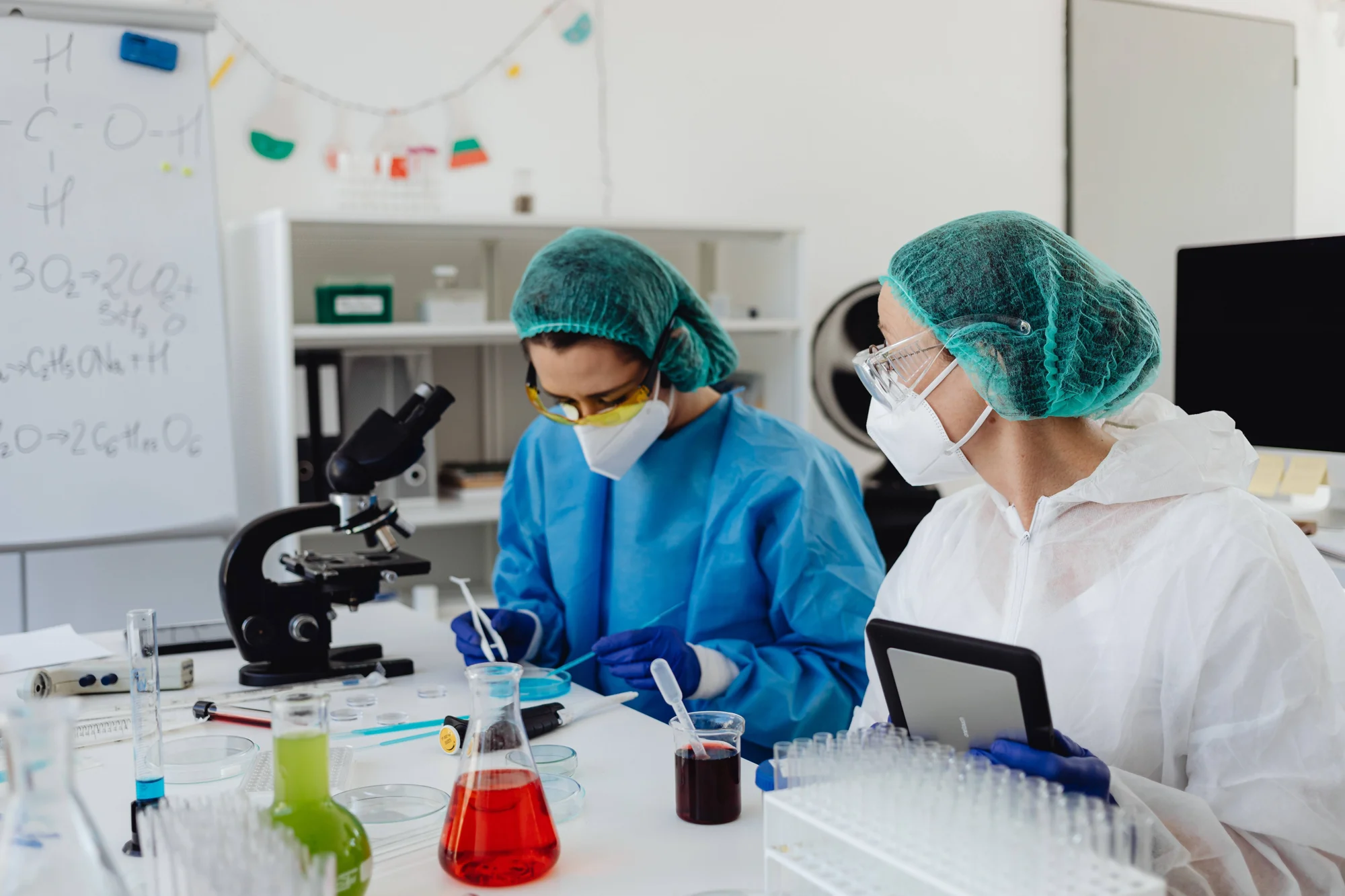 Dos investigadoras con equipo de protección manipulan muestras junto a un microscopio y matraces con líquidos de colores en un laboratorio.