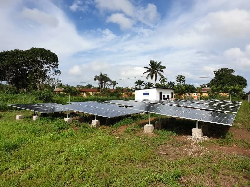 Instalación de paneles solares sobre soportes metálicos en terreno rural con vegetación tropical y una pequeña caseta de control al fondo.