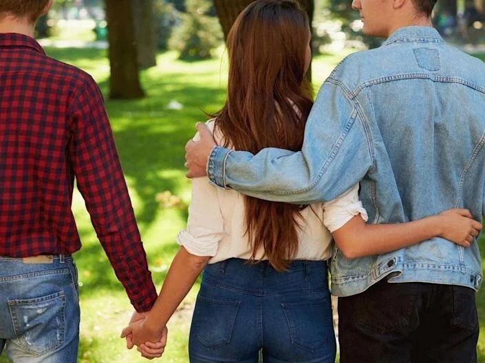 Una mujer camina abrazada por un hombre mientras toma de la mano a otro, simbolizando infidelidad o triángulo amoroso.