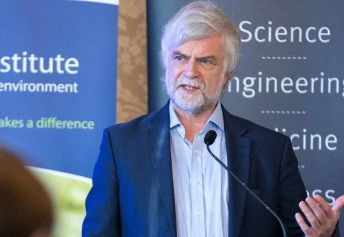 Hombre de cabello canoso y barba blanca habla frente a un micrófono en un evento de ciencia e ingeniería.