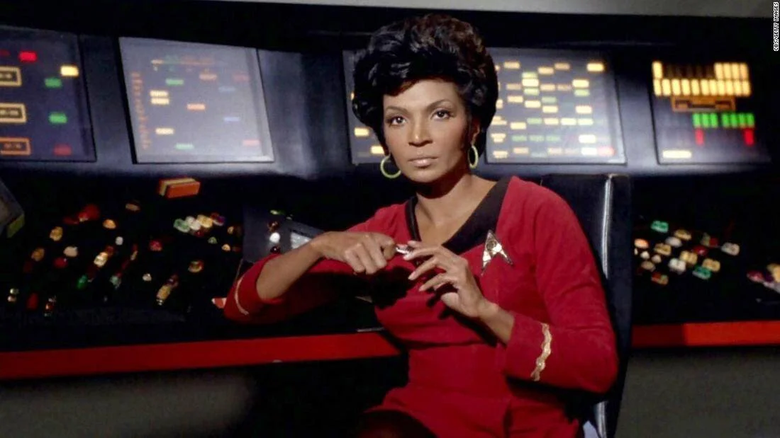 Actriz vestida con uniforme rojo de Star Trek sentada frente al panel de comunicaciones de la Enterprise.