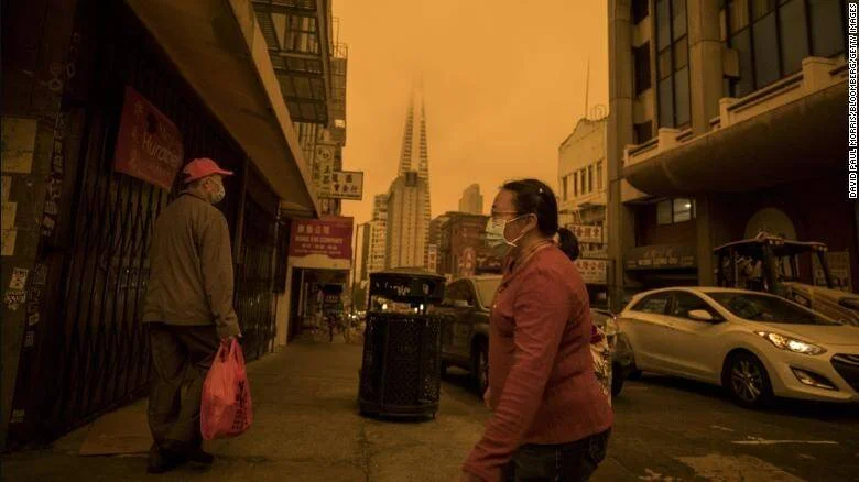 Peatones con mascarilla caminan bajo un cielo teñido de naranja por el humo de incendios forestales en una ciudad de EE. UU.