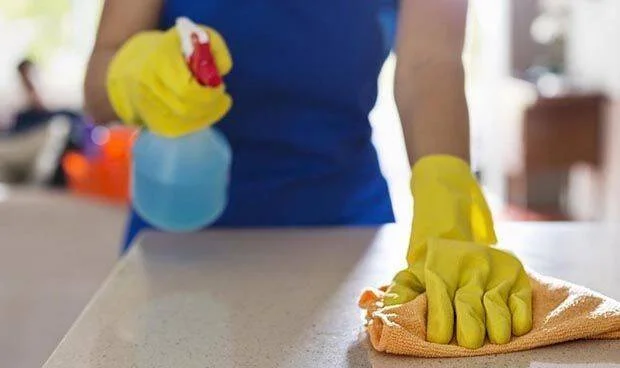 Una persona con guantes amarillos limpia una mesada con un trapo y un pulverizador de producto de limpieza azul.