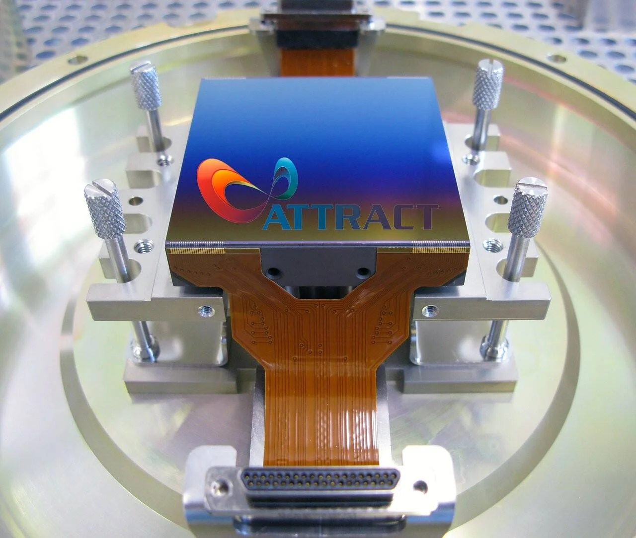 Sensor de detección con logo ATTRACT montado en soporte metálico circular, conectado mediante cable flexible de circuito impreso.
