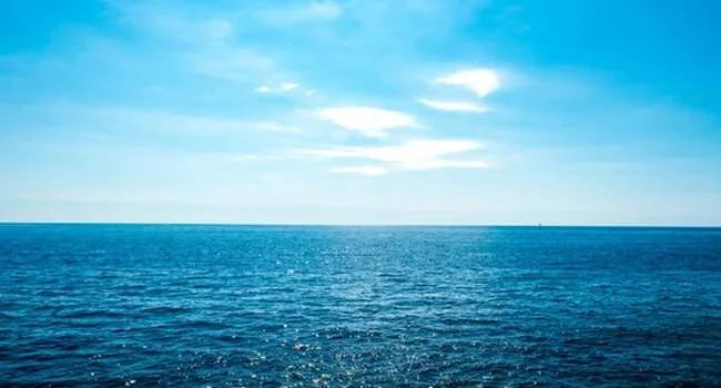 Extensión del océano azul en calma bajo un cielo soleado con nubes dispersas en el horizonte.